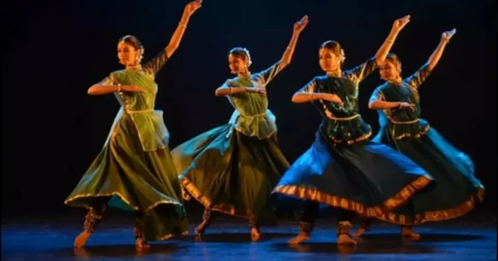 Four classical dancers in green and blue costumes performing on a dark stage